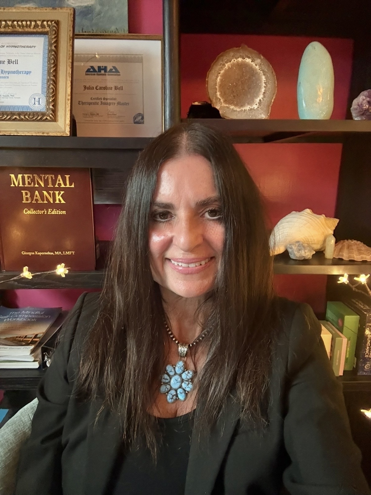 A middle-aged white woman with long brown hair smiles in front of an office full of books and plaques